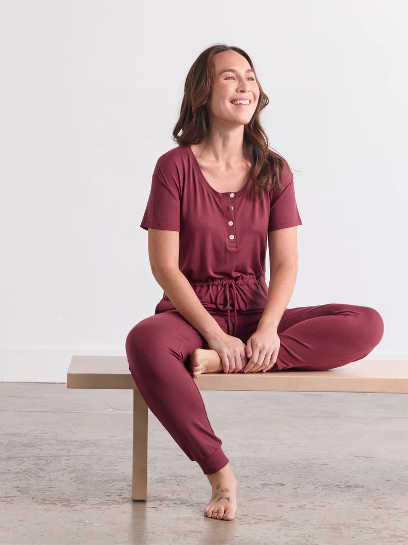 A model seated on a bench wearing an Avocado short sleeve jumpsuit in dark red.